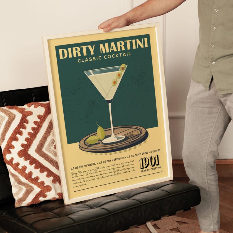 A framed poster of a Dirty Martini cocktail is being held by a person, with a vintage-style design featuring a martini glass and garnishes.