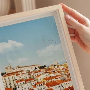 A hand is holding a framed picture of a cityscape with a clear blue sky and fluffy white clouds.