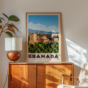 a framed poster of Granada, the capital city of the Alhambra, with a mountainous landscape in the background. The poster is displayed on a wooden cabinet, and there is a lamp and a plant nearby.