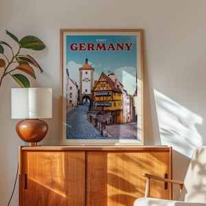 a framed poster of a colorful street scene in Germany, with a clock tower and buildings, hanging on a wall next to a wooden cabinet.