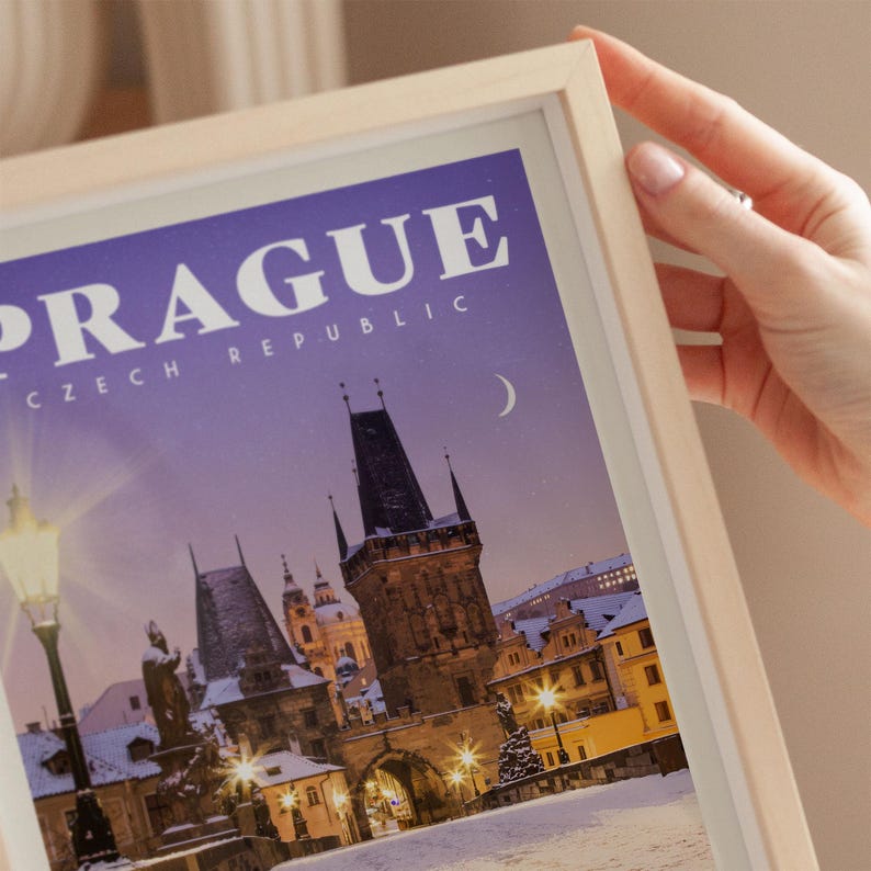 A person's hand is holding a framed picture of Prague, Czech Republic, with a snowy night scene visible.