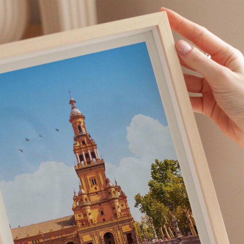 A hand is holding a framed picture of a tall, ornate building with a pointed roof and a clock tower, set against a blue sky with clouds.