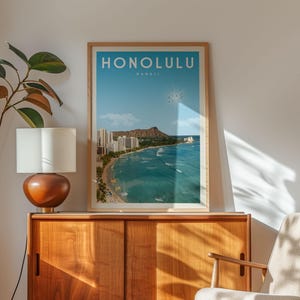 a framed poster of Hawaii, with a view of the ocean and mountains, hanging on a wall next to a lamp and a wooden cabinet.