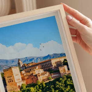 A hand is holding a framed picture of a picturesque cityscape with a castle, mountains, and a clear blue sky in the background.