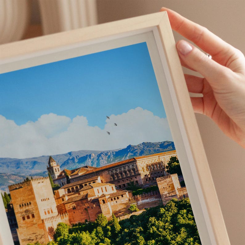 A hand is holding a framed picture of a picturesque cityscape with a castle, mountains, and a clear blue sky in the background.