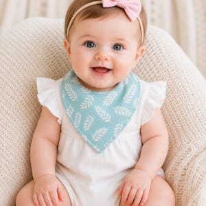 Puede incluir: Un bebé con un body blanco y un babero azul claro con un estampado de hojas blancas. El bebé tiene una diadema con lazo rosa y está sentado en un cojín color crema. El bebé está sonriendo.