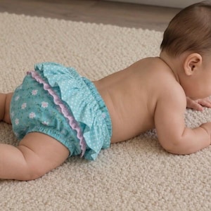 Può includere: Un bambino con un pannolino blu con un motivo floreale e pois bianchi, rifinito con un bordo a balze rosa. Il bambino è sdraiato su un tappeto colorato chiaro.