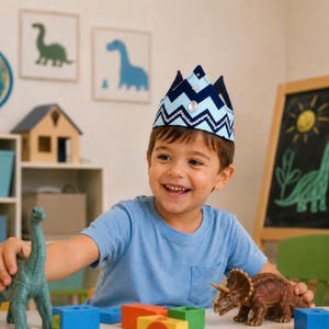 May include: A young child wearing a blue and white crown smiles while playing with dinosaur toys. The child is wearing a blue t-shirt and is surrounded by colorful blocks and a chalkboard with a dinosaur drawing.