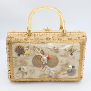 Könnte beinhalten: Eine Vintage-Handtasche aus Korbgeflecht mit einem transparenten Griff und goldfarbener Hardware. Die Frontplatte zeigt eine Ausstellung von Muscheln, Seesternen und anderen Meereselementen, die ein Küstenmotiv schaffen. Die Tasche ist rechteckig und hat einen Schnappverschluss.