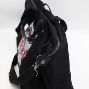 May include: A black canvas duffel bag featuring a central panel with a geometric pattern in red, white, and blue. The bag includes two top handles, a detachable shoulder strap with metal clips, and a zippered closure along the top edge.