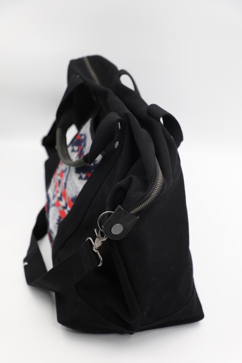 May include: A black canvas duffel bag featuring a central panel with a geometric pattern in red, white, and blue. The bag includes two top handles, a detachable shoulder strap with metal clips, and a zippered closure along the top edge.
