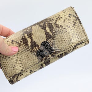 May include: A rectangular wallet featuring a beige and dark brown snakeskin pattern. The front flap is secured with a circular silver-toned metal clasp that displays the text "KENZO" in a repeating circular arrangement around the center.