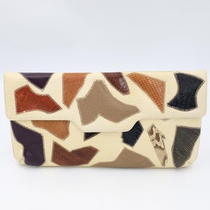 May include: Cream-colored clutch with a patchwork design. The patches are various shapes and colors, including brown, tan, black, and purple. The clutch has a fold-over flap and a rectangular shape.