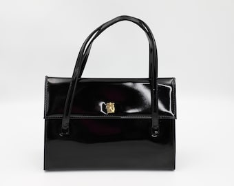 Puede incluir: Un bolso de mano de charol negro con cierre dorado. El bolso tiene un asa superior y un cierre de solapa.
