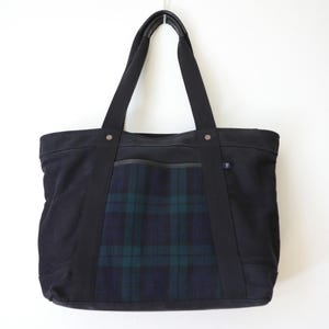 Puede incluir: Un bolso tote negro que presenta un panel central con un patr&oacute;n de cuadros en verde oscuro y azul marino. El bolso tiene dos asas de tela negra, una abertura horizontal superior y una peque&ntilde;a etiqueta rectangular en el lateral del panel.