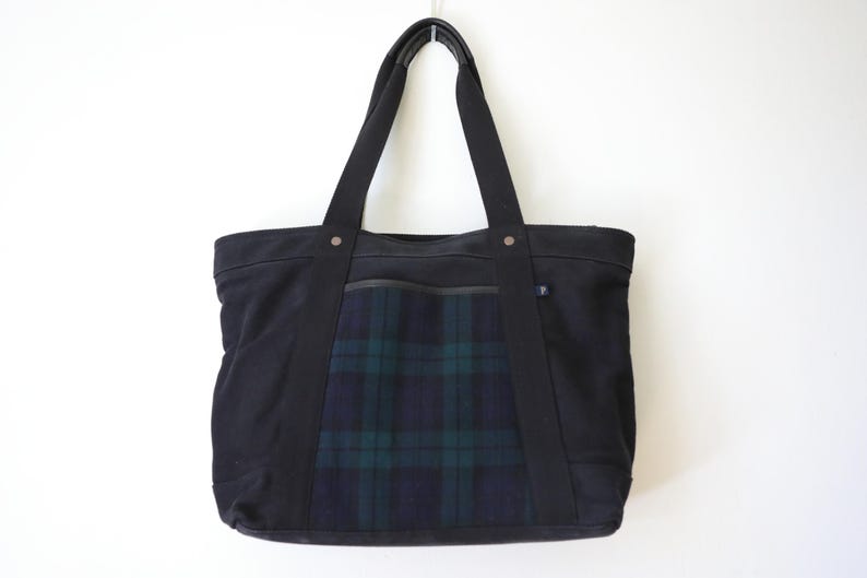 Puede incluir: Un bolso tote negro que presenta un panel central con un patr&oacute;n de cuadros en verde oscuro y azul marino. El bolso tiene dos asas de tela negra, una abertura horizontal superior y una peque&ntilde;a etiqueta rectangular en el lateral del panel.