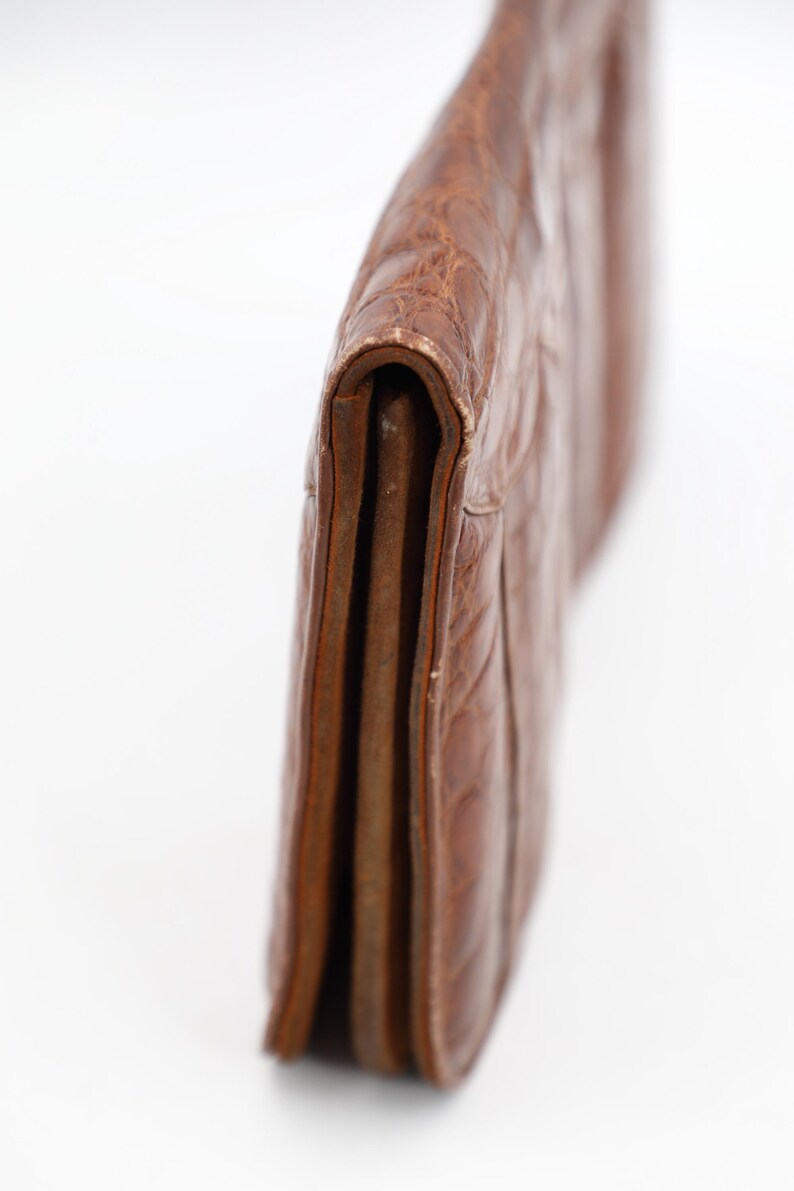 May include: A brown leather clutch bag with a textured, embossed pattern. The bag is folded over, revealing the interior layers. The leather has a worn appearance. The design is simple and classic, suitable for various occasions.