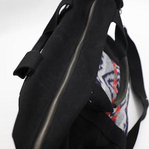 May include: A black duffel bag featuring a central zipper closure, two sturdy black handles, and a side panel decorated with a grey, white, and red geometric pattern. The bag is constructed from a textured fabric and includes a black shoulder strap.