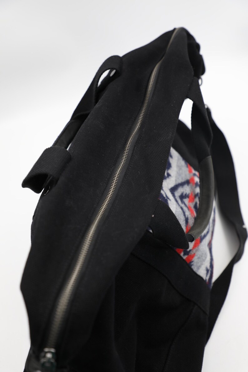 May include: A black duffel bag featuring a central zipper closure, two sturdy black handles, and a side panel decorated with a grey, white, and red geometric pattern. The bag is constructed from a textured fabric and includes a black shoulder strap.