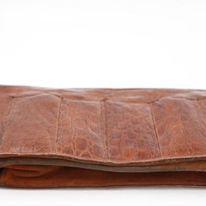 May include: A brown leather clutch bag with a textured, crocodile-like pattern. The rectangular bag has a folded design and a contrasting suede trim. The bag is set against a white background.