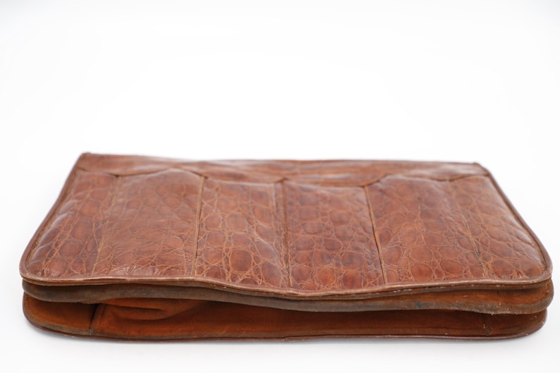 May include: A brown leather clutch bag with a textured, crocodile-like pattern. The rectangular bag has a folded design and a contrasting suede trim. The bag is set against a white background.