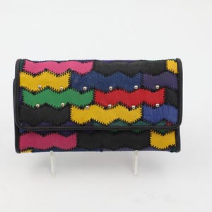 May include: A rectangular wallet featuring a multi-colored patchwork design with wavy, zigzag-stitched edges. The wallet displays alternating blocks of pink, yellow, green, blue, red, and black leather, accented with small silver-toned metal studs.