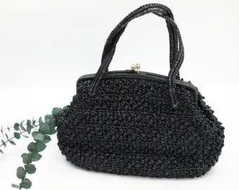 May include: A black handbag with a textured, crocheted surface and a structured frame. The bag features two braided handles and a small, metallic kiss-lock closure at the top. The design is compact with a rounded base and a firm, arched opening.