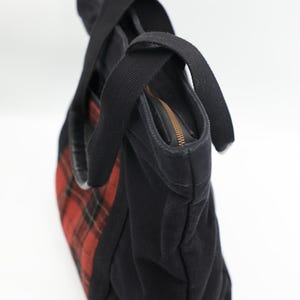 Puede incluir: Un bolso tote de lona negra con un patr&oacute;n de cuadros rojos y negros en el panel lateral. El bolso incluye dos asas de tela negra y un cierre de cremallera superior con un riel met&aacute;lico, proporcionando una apertura segura al interior.
