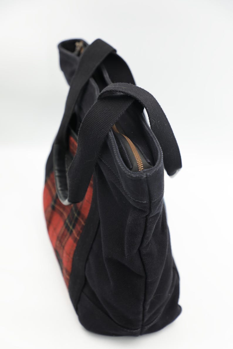 Puede incluir: Un bolso tote de lona negra con un patr&oacute;n de cuadros rojos y negros en el panel lateral. El bolso incluye dos asas de tela negra y un cierre de cremallera superior con un riel met&aacute;lico, proporcionando una apertura segura al interior.