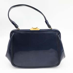 Puede incluir: Un bolso vintage azul marino con un acabado brillante. El bolso presenta una forma rectangular estructurada, un asa curva y un cierre dorado. El asa está sujeta con pequeños herrajes dorados.