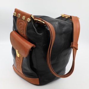 May include: A black leather bucket bag featuring tan leather accents, a front pocket with a gold-tone clasp, and a tan shoulder strap. The bag includes a vertical tan leather panel with embossed circular logos and four gold-tone metal studs at the top.