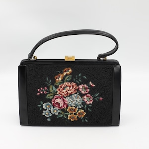Pode incluir: Bolsa preta com textura de contas e alça superior. A frente da bolsa apresenta um design floral bordado em tons de rosa, azul e amarelo. O fecho é dourado.