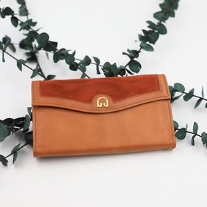 May include: A tan leather clutch with a rust-colored suede flap and a gold-tone clasp. The rectangular wallet is set against a white background with green eucalyptus branches.