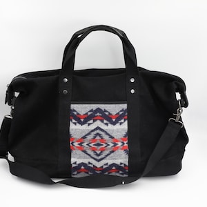 May include: A black duffel bag featuring two top handles and a detachable shoulder strap. The center panel displays a gray, navy blue, and red geometric pattern. The bag includes silver-toned metal rivets securing the handles to the main body.