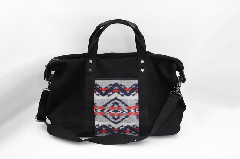 May include: A black duffel bag featuring two top handles and a detachable shoulder strap. The center panel displays a gray, navy blue, and red geometric pattern. The bag includes silver-toned metal rivets securing the handles to the main body.