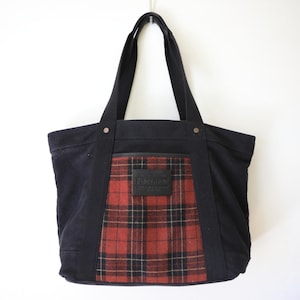 Puede incluir: Un bolso tote de lona negra con un panel central de estampado de cuadros en rojo, negro y blanco. El bolso incluye dos correas negras para el hombro, remaches met&aacute;licos plateados en la base de las correas y un parche rectangular de cuero.