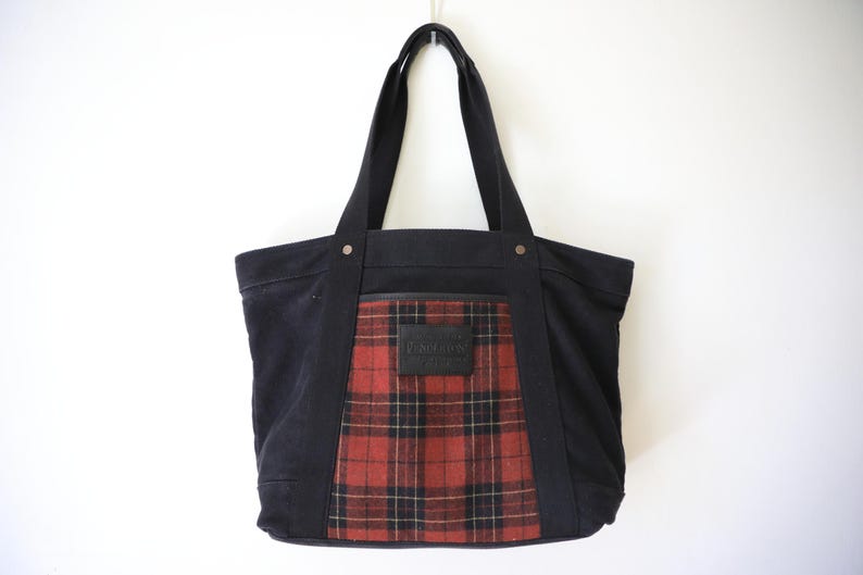Puede incluir: Un bolso tote de lona negra con un panel central de estampado de cuadros en rojo, negro y blanco. El bolso incluye dos correas negras para el hombro, remaches met&aacute;licos plateados en la base de las correas y un parche rectangular de cuero.