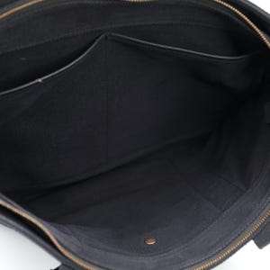 Puede incluir: Un bolso tote de lona negra con interior abierto que cuenta con un compartimento central con cremallera, un bolsillo lateral y un peque&ntilde;o bot&oacute;n de presi&oacute;n met&aacute;lico en la base. El bolso incluye un tirador de cremallera de lat&oacute;n y asas de tela.
