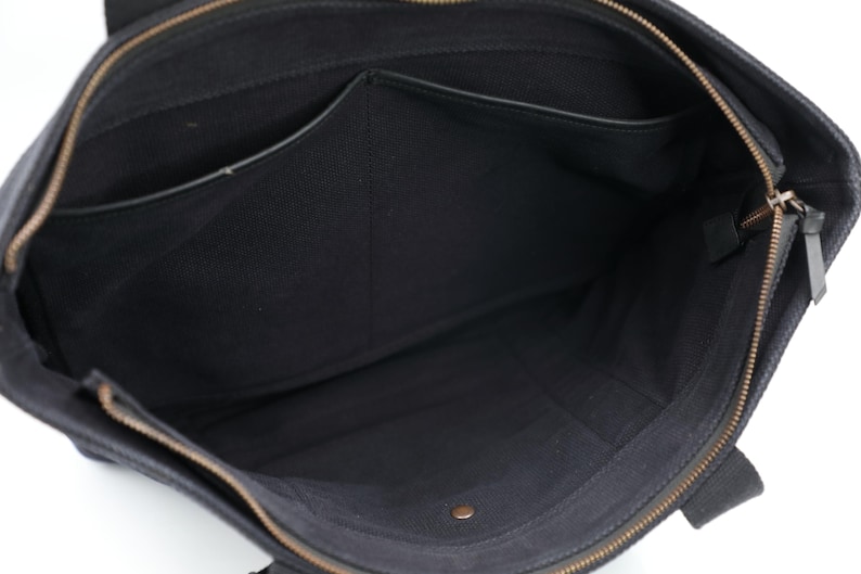Puede incluir: Un bolso tote de lona negra con interior abierto que cuenta con un compartimento central con cremallera, un bolsillo lateral y un peque&ntilde;o bot&oacute;n de presi&oacute;n met&aacute;lico en la base. El bolso incluye un tirador de cremallera de lat&oacute;n y asas de tela.