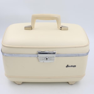 May include: A cream-colored hard-shell vanity case with a top handle, a silver-toned metal trim, and a central locking latch. The front features the word Samsonite in black script lettering. The case stands on four small feet at the base.