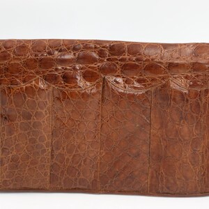 May include: A brown leather clutch bag with a crocodile skin texture. The rectangular bag has a flap closure and a rounded bottom. The leather is a warm brown colour with tone variations, and the surface has a scale-like pattern.