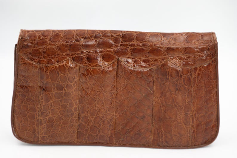 May include: A brown leather clutch bag with a crocodile skin texture. The rectangular bag has a flap closure and a rounded bottom. The leather is a warm brown colour with tone variations, and the surface has a scale-like pattern.