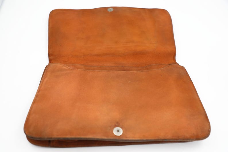 May include: An open, orange-brown leather handbag. The bag has a rectangular shape with a flap closure secured by a silver snap. The interior is visible, showing the soft, suede-like texture of the leather. The bag is likely vintage.