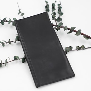May include: A black leather wallet, rectangular in shape, resting on a white surface. Sprigs of greenery are arranged around the wallet. The wallet is closed and has a smooth texture, with a simple design.