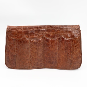 May include: A brown leather clutch bag with a crocodile skin effect. The rectangular bag features a scalloped flap and a smooth, glossy finish. The bag is a medium size and is set against a white background.