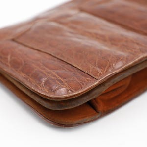 May include: Close-up of a brown leather clutch with a textured, crocodile-like pattern. The clutch has a rectangular shape with rounded edges and a visible seam. The interior appears to be a lighter shade of brown.