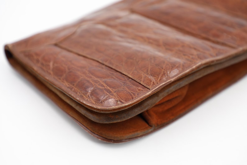 May include: Close-up of a brown leather clutch with a textured, crocodile-like pattern. The clutch has a rectangular shape with rounded edges and a visible seam. The interior appears to be a lighter shade of brown.