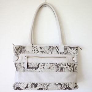 May include: Cream-colored tote bag with a snakeskin pattern. The bag features a rectangular shape with a zippered top and a front zippered pocket. The handles are long and made of the same cream-colored material.