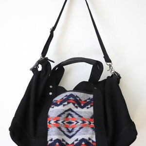 May include: A black shoulder bag featuring a central panel with a grey, navy blue, and orange geometric pattern. The bag includes two black top handles, a detachable black shoulder strap with metal hardware, and two silver-tone rivets.