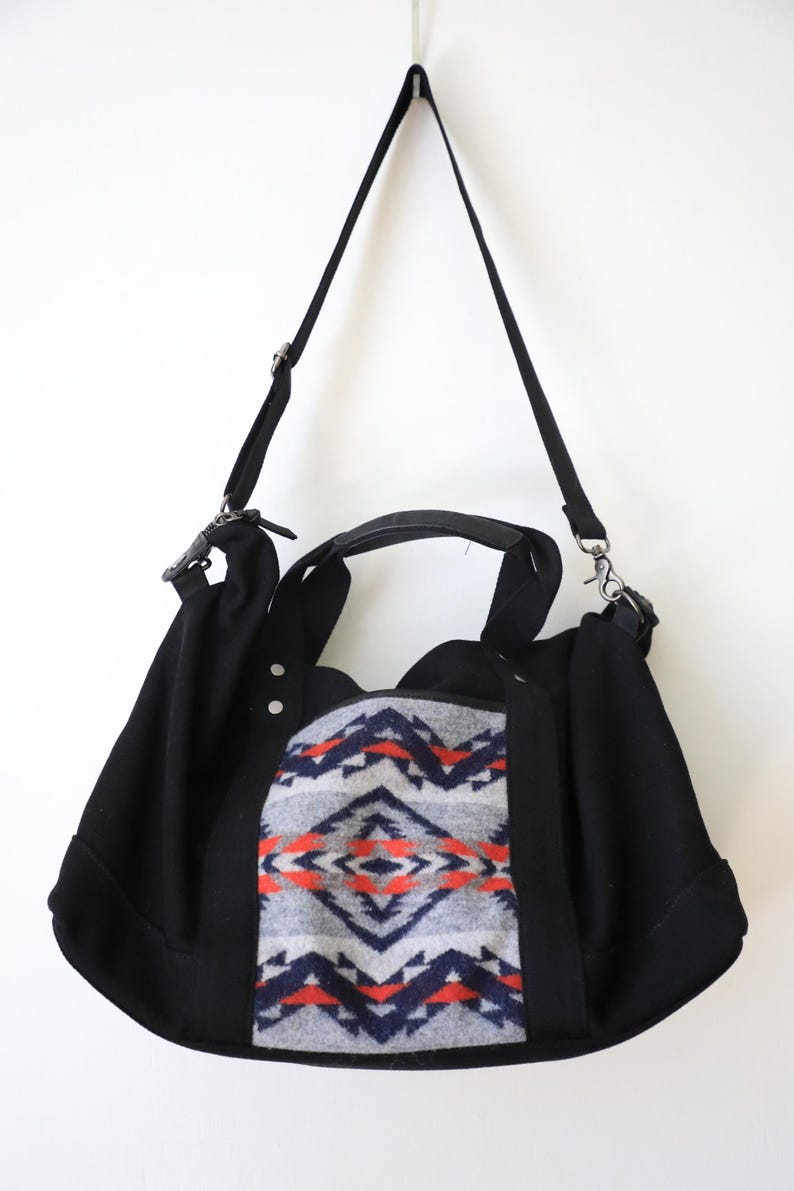 May include: A black shoulder bag featuring a central panel with a grey, navy blue, and orange geometric pattern. The bag includes two black top handles, a detachable black shoulder strap with metal hardware, and two silver-tone rivets.